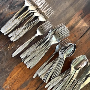 May include: A collection of stainless steel flatware including multiple forks and spoons. Each piece features a polished finish and a decorative embossed floral pattern along the handle. The utensils are arranged in groups.