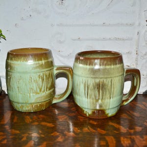 May include: Two ceramic mugs shaped like wooden barrels with a light green and brown drip glaze finish. Each mug features a sturdy side handle and a cylindrical body with horizontal ridges mimicking barrel staves, showcasing a glossy texture.