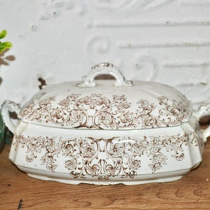 May include: A white and brown antique serving dish with a lid. The dish has a floral pattern and two handles on the sides. The lid has a handle on top. The dish is on a wooden surface.