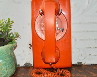 Puede incluir: Un teléfono de pared con disco giratorio de color naranja brillante con un auricular a juego que descansa en la base central. El dial circular presenta números y letras blancos, y un cable naranja en espiral sale de la base rectangular.