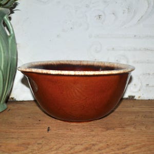 May include: A brown ceramic mixing bowl with a white rim. The bowl has a rounded shape with a slightly flared rim. The interior of the bowl is a darker brown color.