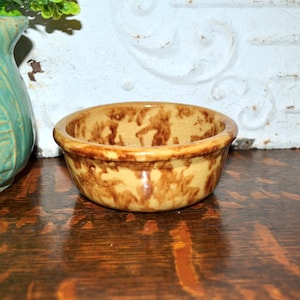 May include: A small, round ceramic bowl featuring a mottled, sponge-painted pattern in shades of tan and dark brown. The bowl has a smooth, glossy finish and a thick, rounded rim, presenting a consistent, speckled appearance across its exterior surface.