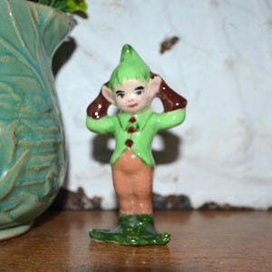 May include: A ceramic elf figurine featuring a pointed green hat, a light green jacket with three dark red buttons, and tan trousers. The elf has brown pointed ears and is depicted with both hands raised to the sides of its head in a standing pose.