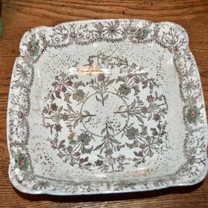 May include: Square, antique white ceramic dish with a floral design in muted green, pink, and gold. The rim has a decorative edge. The dish is on a wooden surface.