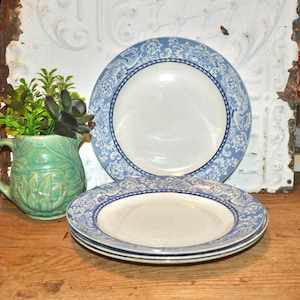 May include: A stack of three white and blue patterned plates with a floral design. The plates are stacked on a wooden surface. A green pitcher with a plant is to the left of the plates.