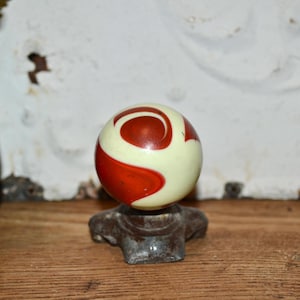 Puede incluir: Un pomo de armario vintage con una bola de color crema con un diseño de remolino rojo. El pomo está montado sobre una base de metal oscuro y envejecido, mostrando una estética clásica y retro.
