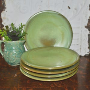 May include: A stack of four round ceramic plates in a muted sage green glaze with subtle brown undertones. Beside the plates sits a matching green ceramic pitcher containing a small artificial succulent plant with dark green leaves.