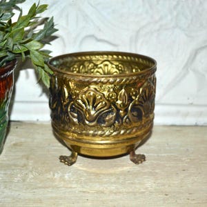 May include: A small, antique brass planter with ornate floral and leaf detailing. The planter has a round shape, three small feet, and a decorative rope-like trim around the top. The brass has a warm, aged patina.