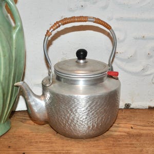 Puede incluir: Una tetera vintage de color plateado con una superficie texturizada. Cuenta con un asa de metal curvada con un agarre marrón tejido, un pomo negro en la tapa y una pequeña palanca roja. La tetera está diseñada para su uso en la estufa.