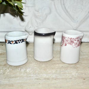 May include: Three small, white ceramic pitchers. One has a black and orange patterned band, another has a black rim, and the third has a floral design in red. They are arranged on a light-colored wooden surface.