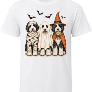 May include: A white short-sleeved t-shirt featuring a graphic of three black and white dogs dressed for Halloween. One dog is wrapped in mummy bandages, one is wearing a white ghost sheet, and one wears an orange witch hat and cape with bats.