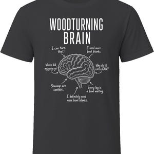 May include: A charcoal gray t-shirt featuring a white graphic of a human brain. The text reads WOODTURNING BRAIN, with arrows pointing to various parts of the brain labeled with phrases like I can turn that, I need more bowl blanks, and Shavings are confetti.