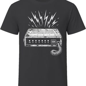 May include: A charcoal gray t-shirt featuring a white graphic print of a vintage 56K modem. The design includes lightning bolts above the device, status indicator lights labeled PWR, CO, RD, SD, TR, OH, AA, and a coiled telephone cord extending downward.