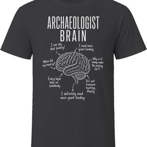 May include: A dark grey t-shirt featuring a white graphic of a human brain. The text reads ARCHAEOLOGIST BRAIN, with arrows pointing to various parts of the brain labelled with phrases like I can dig that history, I need more grant funding, and Where did my trowel go?