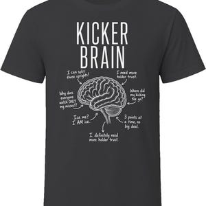 May include: A charcoal grey short-sleeve t-shirt featuring a white graphic of a human brain. The text reads KICKER BRAIN with various labels pointing to parts of the brain, including I can split those uprights, I need more holder trust, and 3 points at a time, no big deal.