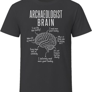 Peut inclure: Un t-shirt gris foncé avec un graphique blanc représentant un cerveau humain. Le texte indique ARCHAEOLOGIST BRAIN, avec des flèches pointant vers diverses parties du cerveau accompagnées de phrases comme I can dig that history et I need more grant funding.