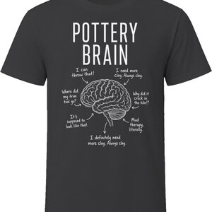 May include: A charcoal gray short-sleeve t-shirt featuring a white graphic of a human brain. The text POTTERY BRAIN is printed above the brain, with arrows pointing to various parts labeled with phrases like I can throw that and Mud therapy.