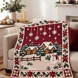 Puede incluir: Una manta de ganchillo rectangular que presenta una escena de pueblo invernal con casas nevadas, un muñeco de nieve y copos de nieve sobre un fondo rojo intenso, enmarcada por un borde decorativo de flores de pascua rojas y hojas de acebo.