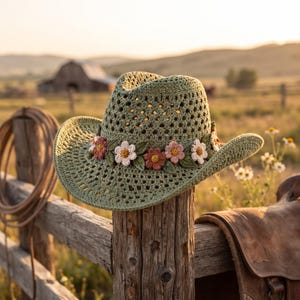 Peut inclure: Un chapeau de cowboy en crochet vert sauge avec un motif ajouré et une bande décorative composée de petites fleurs en crochet multicolores et de feuilles vertes entourant la calotte. Le chapeau présente un large bord incurvé et un sommet pointu.