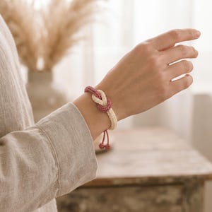 May include: A woven cord bracelet featuring a central knot design in cream and dusty rose colours. The bracelet has an adjustable closure with two small dangling cord ends, each finished with a matching dusty rose spherical bead at the very tip.