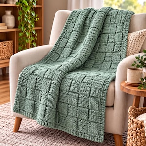 May include: A sage green knitted throw blanket featuring a prominent basketweave texture. The blanket is draped over a light-colored upholstered armchair, showcasing thick yarn loops and a consistent grid-like pattern across the entire surface.