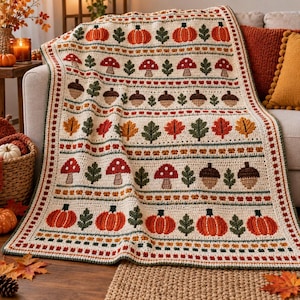 May include: A cream-colored crochet throw blanket featuring horizontal rows of autumn-themed motifs including orange pumpkins, red mushrooms, brown acorns, and green leaves. The blanket has a decorative border and a textured, handmade appearance.