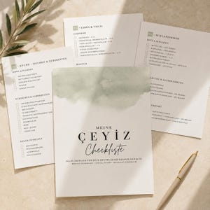 Op de afbeelding: Een set witte papieren checklists met zwarte tekst en een saliegroen aquarelaccent. De centrale kaart bevat de tekst 'MEINE ÇEYIZ Checkliste' en 'ALLES IM BLICK FÜR DEIN ERSTES GEMEINSAMES ZUHAUSE'. Een beige pen ligt naast de kaarten.