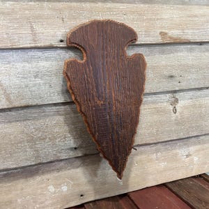 May include: A dark brown wooden wall plaque shaped like a large arrowhead. The wood features a prominent grain pattern with a textured, weathered surface and a pointed bottom edge, while the top section has two symmetrical side notches.