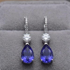 May include: A pair of silver-toned drop earrings featuring a small circular clear stone set above a larger pear-shaped deep blue stone. The upper portion of each earring consists of a thin hoop encrusted with small clear stones for added sparkle.