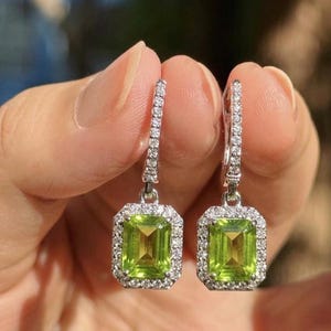 Pode incluir: Um par de brincos pendentes com peridotos verdes de corte esmeralda em metal prateado. Cada pedra central é rodeada por um halo de pequenas pedras brancas redondas, com pedras brancas adicionais ao longo dos ganchos verticais dos brincos.