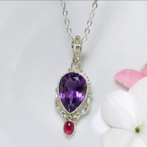 Könnte beinhalten: Eine silberne Anhängerkette mit einem großen, birnenförmigen, facettierten violetten Amethyst in einer dekorativen Silberfassung, wobei sich ein kleiner, runder, im Cabochon-Schliff gehaltener roter Rubin unter dem Amethyst befindet.