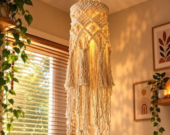 May include: A cylindrical macrame pendant light fixture featuring an intricate knotted diamond pattern, a central light source, and long, flowing fringe tassels hanging from the base. The off-white cord construction creates a textured appearance.