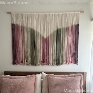 May include: A wall hanging made of cream-colored cotton cord suspended from a wooden dowel. The lower portion of the hanging features a V-shaped dip-dyed pattern in shades of dusty rose, sage green, and deep plum, creating a layered color effect.