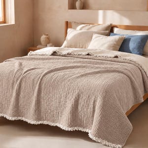 Op de afbeelding: Een beige, getextureerde sprei met een franjerand gedrapeerd over een bed. Het bed is voorzien van twee beige kussens, één wit kussen en één blauw kussen, die tegen een houten hoofdeinde rusten in een kamer met lichtgekleurde muren.