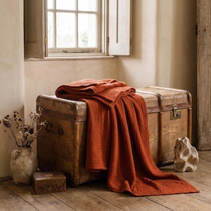 May include: A textured, rust-colored muslin blanket draped over a large, weathered wooden storage trunk with metal latches. The blanket features a soft, crinkled fabric finish and hangs down the front of the rectangular chest in deep, loose folds.