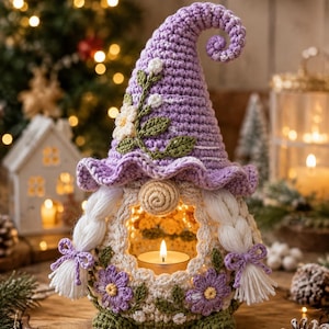 Könnte beinhalten: Eine gehäkelte Wichtelfigur mit einer hohen, spitzen lavendelfarbenen Mütze, die mit weißen Blüten und grünen Blättern verziert ist. Der Wichtel hat weißes geflochtenes Haar, eine beige Nase und eine hohle Mitte mit einem Teelicht, verziert mit lila Blüten.