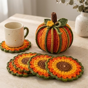May include: A crocheted pumpkin in orange and red with green ribbed accents, a brown stem, and a small yellow flower. Beside it are four round, sunflower-patterned crochet coasters in shades of brown, yellow, orange, and green, with one under a mug.