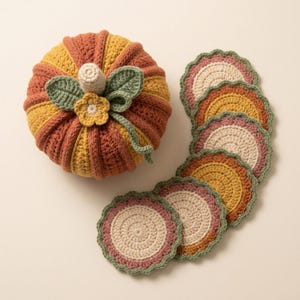 May include: A crocheted pumpkin in shades of mustard yellow and burnt orange with a cream stem, green leaves, and a small yellow flower, accompanied by a set of six circular crocheted coasters in matching colors with scalloped green edges.