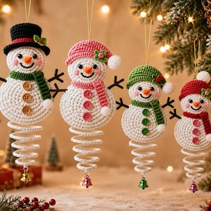 May include: Four crocheted snowman ornaments hanging by gold strings. Each snowman has a white body, orange nose, and a spiral base with a small bell. They wear colorful hats and scarves, featuring brown, pink, green, and red buttons on their chests.