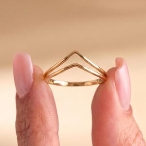 Op de afbeelding: Een goudkleurige ring met een dubbel chevron- of V-vormig ontwerp. De band bestaat uit twee parallelle, dunne, gladde metalen lijnen die samenkomen in een centraal punt, wat zorgt voor een geometrisch silhouet met een gepolijste afwerking.