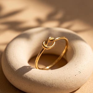 Peut inclure: Une fine bague en or poli présentant un motif de nœud central. L'anneau est circulaire et lisse, arborant une finition métallique réfléchissante qui capte la lumière. La bague est positionnée verticalement, mettant en valeur le détail du nœud.