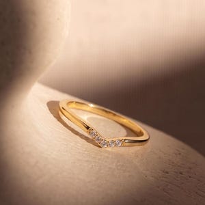 Op de afbeelding: Een gouden ring met een V-vormig chevronontwerp. Het midden van de ring is versierd met vijf kleine, heldere, rondgeslepen stenen die in een rij langs de punt van de V zijn geplaatst, wat zorgt voor een subtiele schittering op het gepolijste goud.