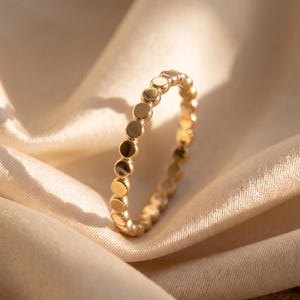 Puede incluir: Un anillo de oro que presenta una banda continua de pequeñas cuentas metálicas circulares y pulidas dispuestas en una sola fila. El anillo tiene un acabado metálico reflectante y un patrón uniforme de esferas conectadas que forman la banda.