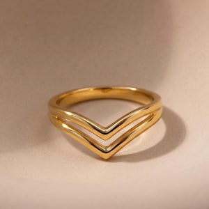 May include: A gold-toned ring featuring a double chevron or V-shaped band design. The ring has a smooth, polished metallic finish with two distinct parallel lines that meet at a central point, creating a symmetrical, angular silhouette.