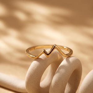 May include: A gold-toned ring featuring a continuous, angular design that mimics the shape of a mountain range or a heartbeat rhythm. The band is smooth and polished, with a distinct peak and valley silhouette that rises from the circular ring base.