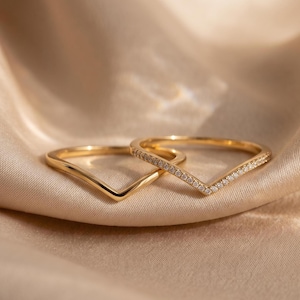 May include: Two gold V-shaped rings resting together. One ring features a smooth, polished gold band, while the other ring is set with a row of small, clear, round-cut stones along the outer edge of the V-shape, creating a matching set of jewellery.