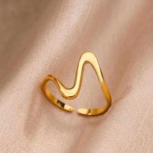 Puede incluir: Un anillo de tono dorado con un diseño ondulado continuo y suave que forma una figura abierta y asimétrica. La banda tiene una superficie pulida y reflectante con una abertura en la parte inferior, lo que indica un ajuste regulable.