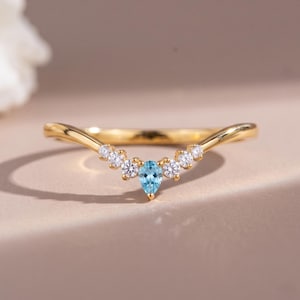 Könnte beinhalten: Ein goldener V-förmiger Ring mit einem zentralen hellblauen Edelstein im Tropfenschliff, der auf jeder Seite von drei kleinen runden weißen Steinen flankiert wird. Die Ringschiene ist poliert und weist eine geschwungene, symmetrische Form auf.