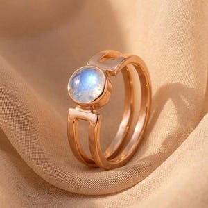 Op de afbeelding: Een roségouden ring met een ronde, iriserende maansteen cabochon in een zetting. De ringband heeft een gespleten ontwerp dat samenkomt in een enkele schacht aan de basis, met een gepolijste metalen afwerking en een glad, afgerond edelsteenoppervlak.