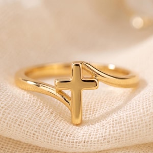 Peut inclure: Une bague dorée polie présentant un motif de croix central. L'anneau possède un design lisse et continu qui se courbe pour s'intégrer à l'élément vertical de la croix, créant une apparence élégante et uniforme avec une finition métallique brillante.